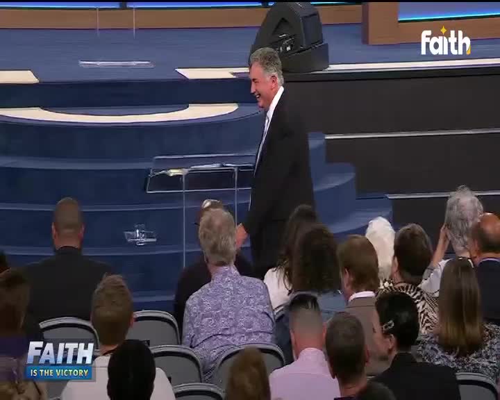 A man in a dark suit, smiling, walks down a blue-carpeted stage, likely on Faith TV in South Africa. The audience watches from rows of grey chairs, some with their backs to the camera.

