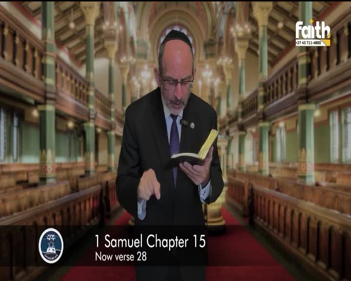 A man in a suit and kippah reads from a small book, his finger pointing to a verse. The background shows the interior of a synagogue, with rows of wooden benches and ornate pillars.