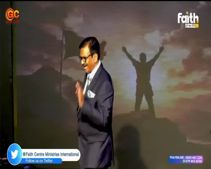 A man in a suit gestures with his hands as he speaks, his image projected onto a screen behind him. The screen displays a silhouette of a person with arms raised in triumph against a dramatic sky, a common scene on Faith TV broadcasts.