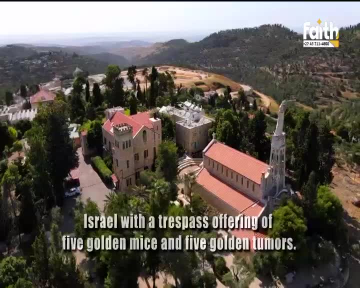 The camera glides over a cluster of buildings nestled into a hillside, their red roofs stark against the green trees. A tall, slender spire rises from one of the structures, catching the sunlight.
