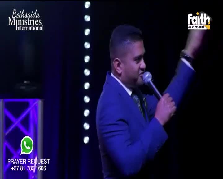 A man in a blue suit speaks into a microphone, his right arm raised. Lights shine behind him, illuminating the stage. A man in a blue suit speaks into a microphone, his right arm raised. Lights shine behind him, illuminating the stage.