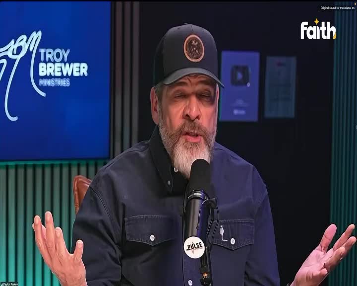 A man with a beard and a baseball cap speaks into a microphone, his hands open and gesturing. Behind him, a screen displays "Troy Brewer Ministries," and the Faith TV logo is visible.