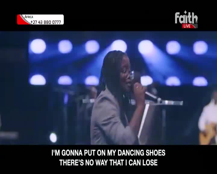 A woman in a grey jacket sings into a microphone on a stage, bathed in blue light. The Faith LIVE broadcast from South Africa shows her passionate performance.