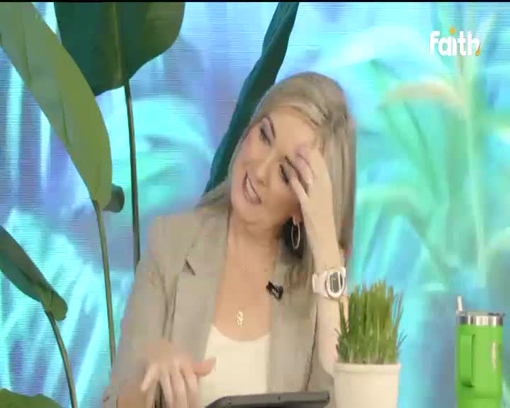 The woman on Faith TV gestures with her hand, her blonde hair falling forward as she touches her forehead. A bright green thermos sits beside a small potted plant on the table.