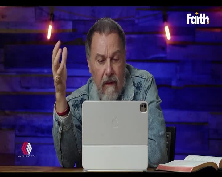 A man with a beard gestures with his hand while looking at a tablet, his denim jacket embroidered with a floral pattern. A Bible rests open on the table beside him, illuminated by the warm glow of stage lights.