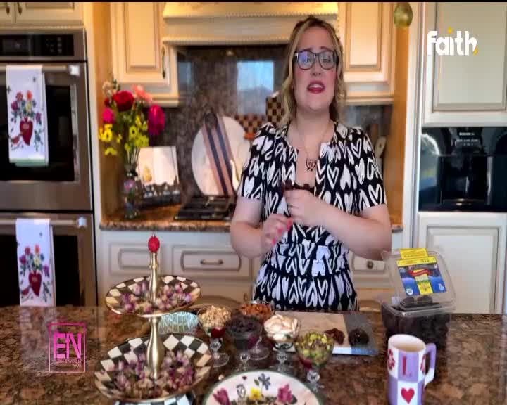 A woman in a black and white patterned dress stands in a kitchen, holding a dark, elongated object. She's preparing ingredients for what looks like a recipe, with bowls of nuts and dried fruit laid out on the counter. The Faith TV logo is visible in the corner, suggesting this is part of a broadcast from South Africa.