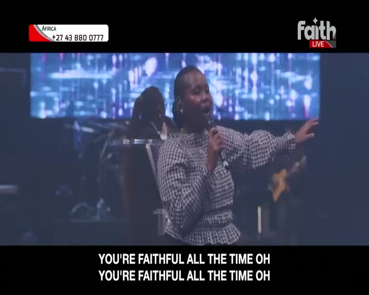 A woman in a checkered top sings passionately into a microphone on a stage in South Africa. The Faith Live broadcast displays lyrics on screen as she gestures with her free hand.