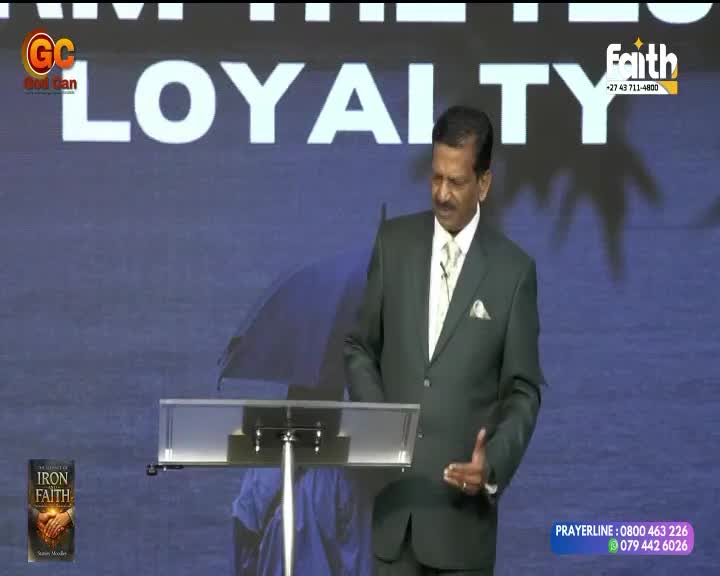 A man in a dark suit stands at a podium, speaking. Behind him, the word "LOYALTY" glows in large white letters on a blue screen. A book titled "Iron Faith" rests on a stand to his left.