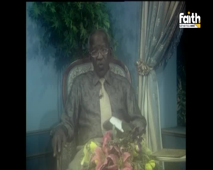 A man in a patterned shirt and tie speaks from a chair, a microphone clipped to his collar. He gestures with a white paper, a bouquet of flowers at his feet.