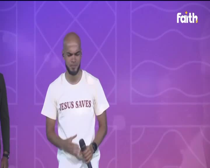 A man with a shaved head stands on a stage, holding a microphone. His white t-shirt bears the words "JESUS SAVES" in dark red lettering. The backdrop is a vibrant purple with a geometric pattern.