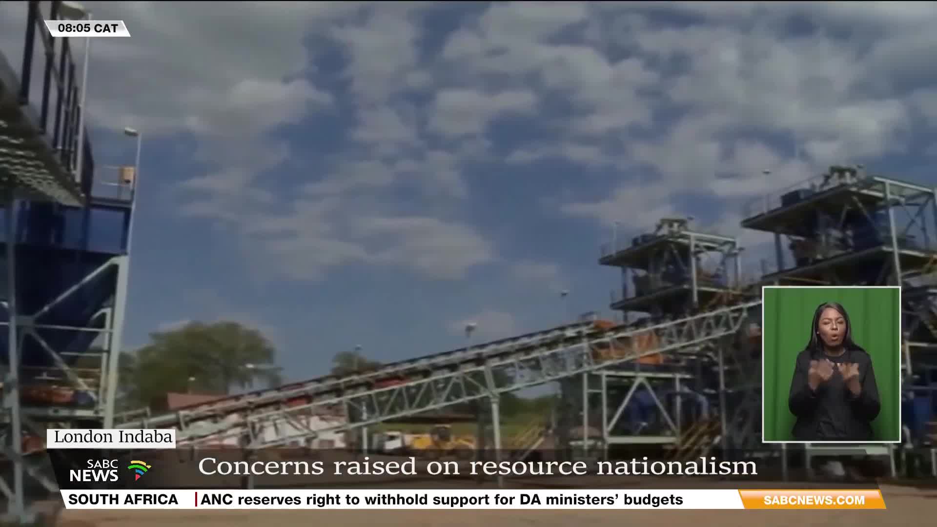 A conveyor belt stretches across the frame, its metal structure against a cloudy sky. The SABC News broadcast from South Africa highlights concerns about resource nationalism.

