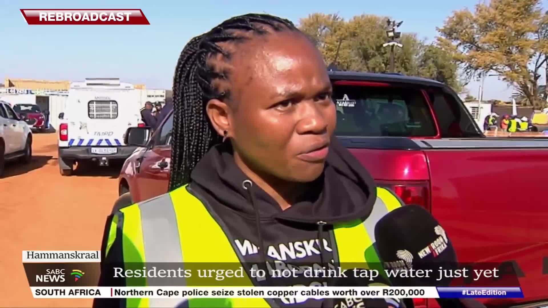 A woman in a reflective vest speaks into a microphone, likely for SABC News, with a red pickup truck behind her. The backdrop includes vehicles and buildings, suggesting a location in South Africa.
