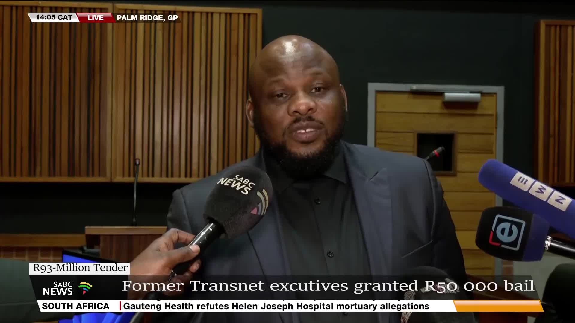A man in a dark suit, speaking into an SABC News microphone, addresses the camera. The live broadcast from Palm Ridge, GP, reports on former Transnet executives granted bail.
