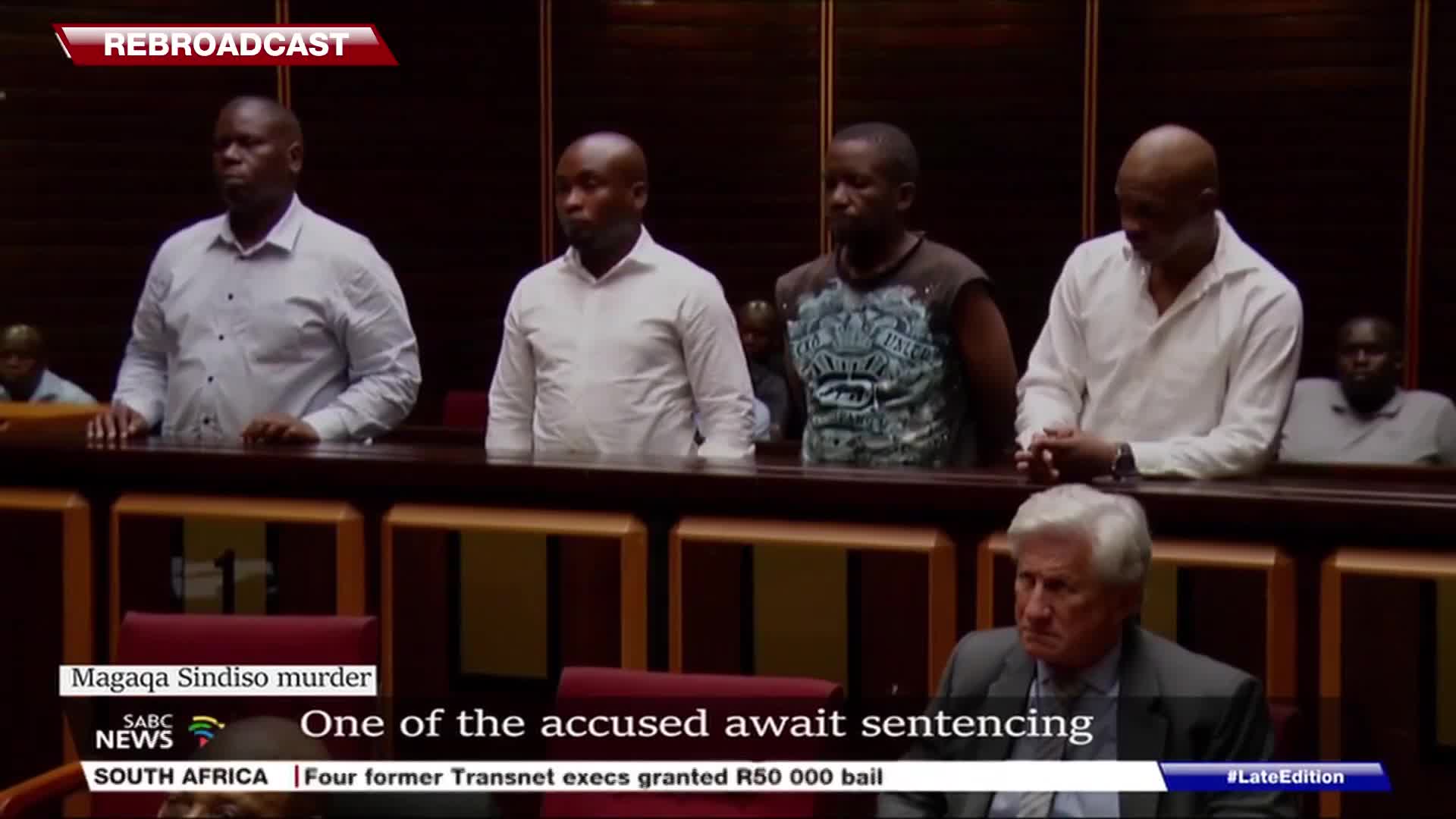Four men stand behind a wooden railing, each in a white shirt except one in a sleeveless top. Below them, a man in a suit sits, looking down, as SABC News reports on the Magaqa Sindiso murder case.
