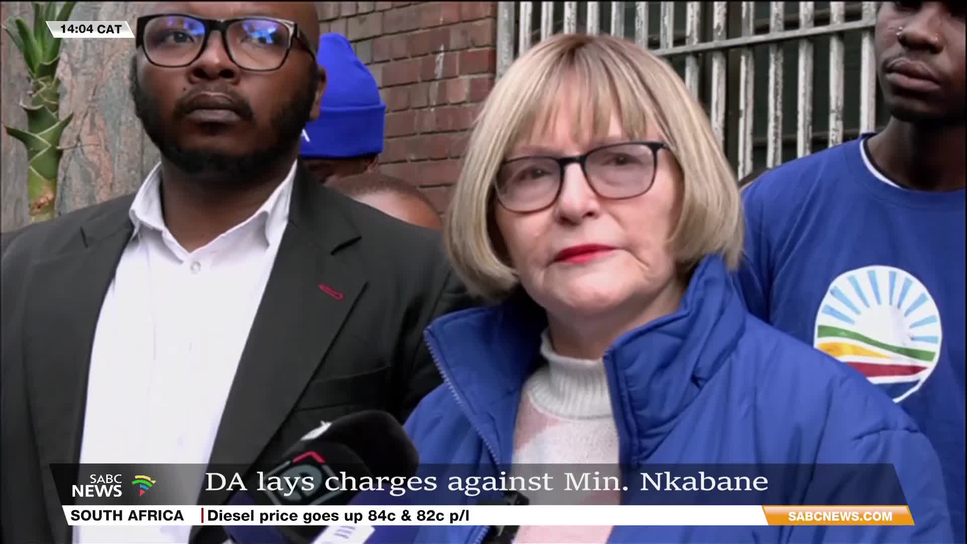 A woman with short blonde hair and glasses speaks into a microphone, flanked by two men. The SABC News graphic at the bottom of the screen reports on the DA laying charges in South Africa.
