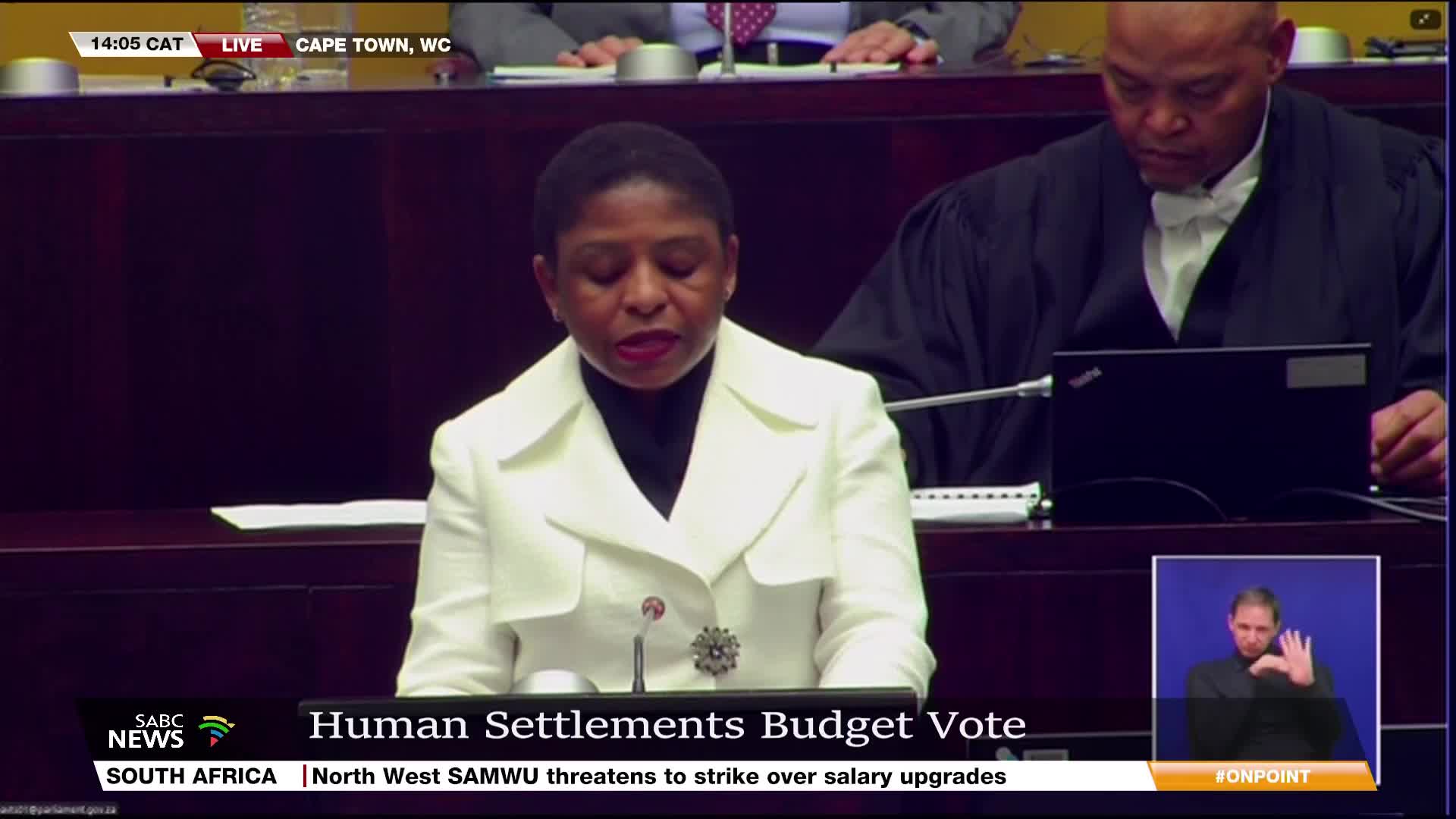 A woman in a white jacket speaks at a podium, the SABC News logo visible in the lower left corner. Behind her, a man in a black robe sits, while a sign language interpreter appears in a small box on the right.
