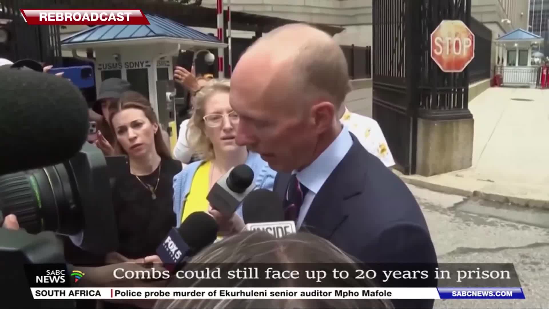 A man in a dark suit speaks into a cluster of microphones, surrounded by reporters and cameras. The SABC News banner at the bottom mentions a police probe in South Africa.
