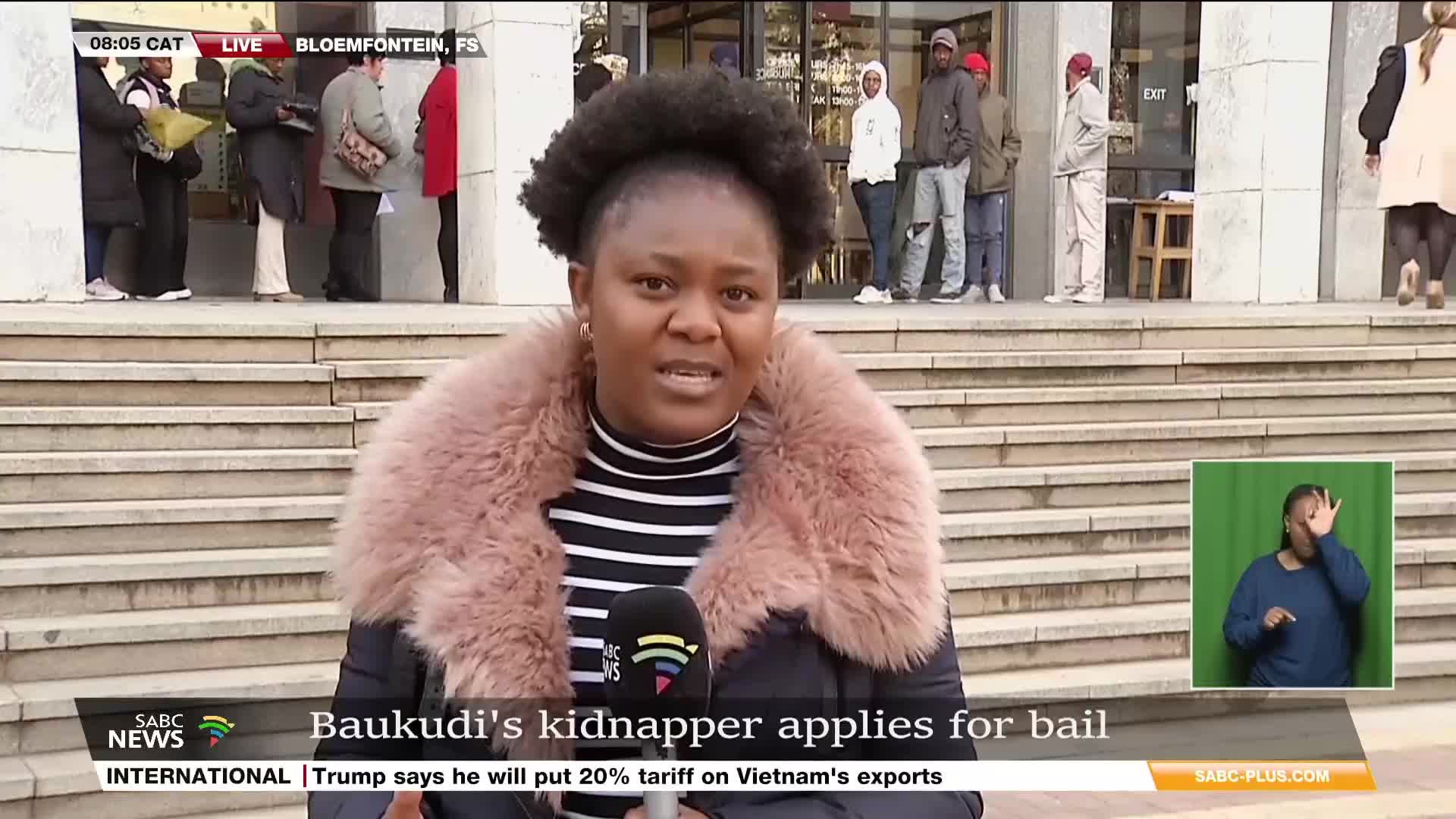 An SABC News reporter, wearing a pink fur-trimmed jacket, speaks directly to the camera in Bloemfontein, South Africa. Behind her, people wait outside a building, while a sign language interpreter appears on a green screen.
