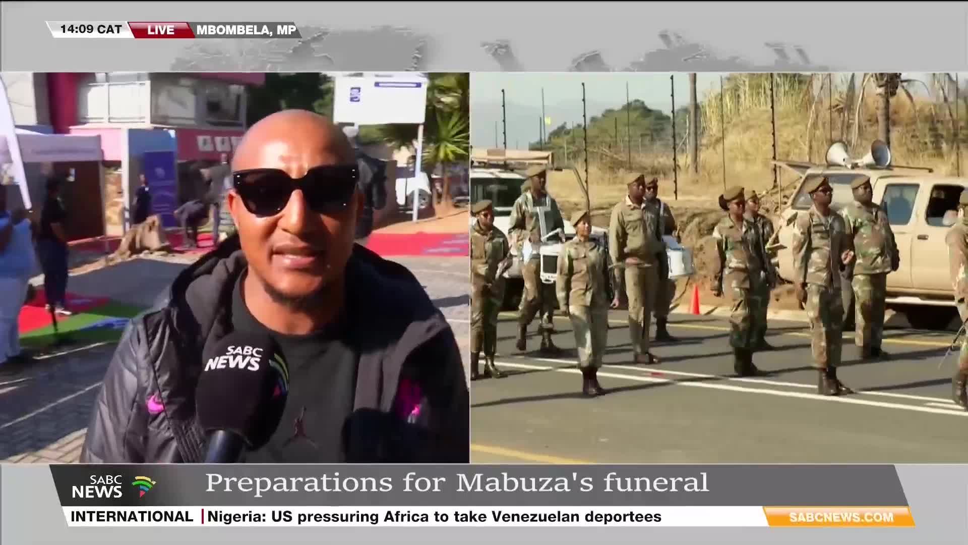 An SABC News reporter is speaking to the camera, while a group of soldiers in camouflage uniforms march in formation on the road behind him. The headline on the screen says "Preparations for Mabuza's funeral" in Mbombela, South Africa.
