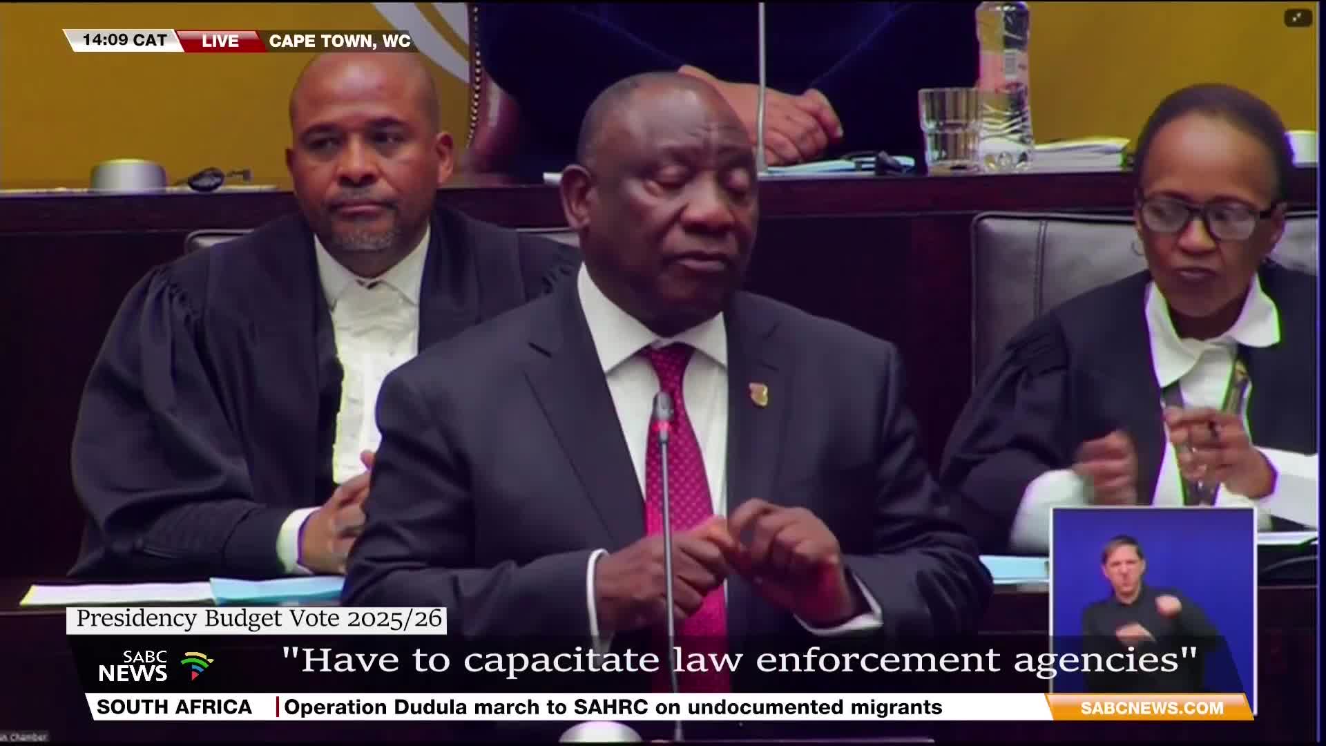 The President of South Africa, wearing a dark suit and a patterned tie, speaks into a microphone. Two other people, dressed in legal robes, sit on either side of him, listening intently.
