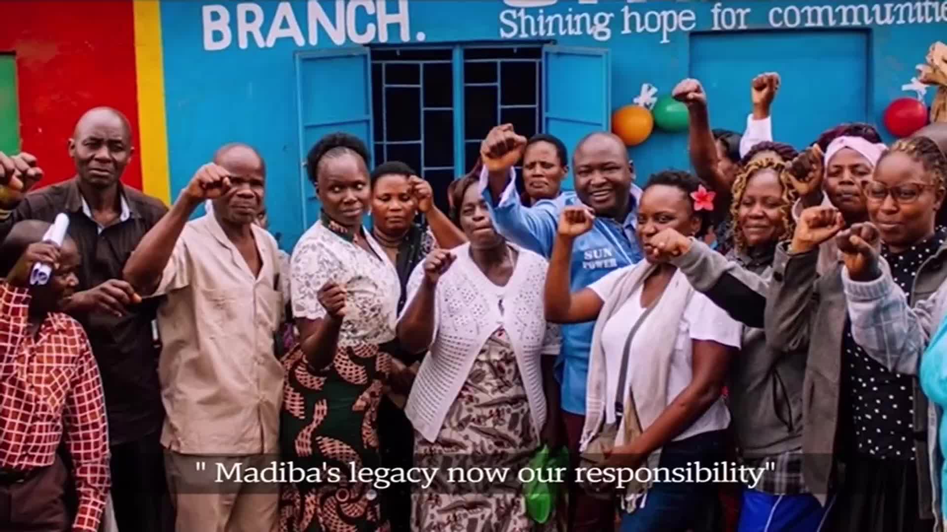 A crowd of people in South Africa, many with their fists raised, stand in front of a building. The words "Madiba's legacy now our responsibility" are displayed below the image, likely from an SABC News report.
