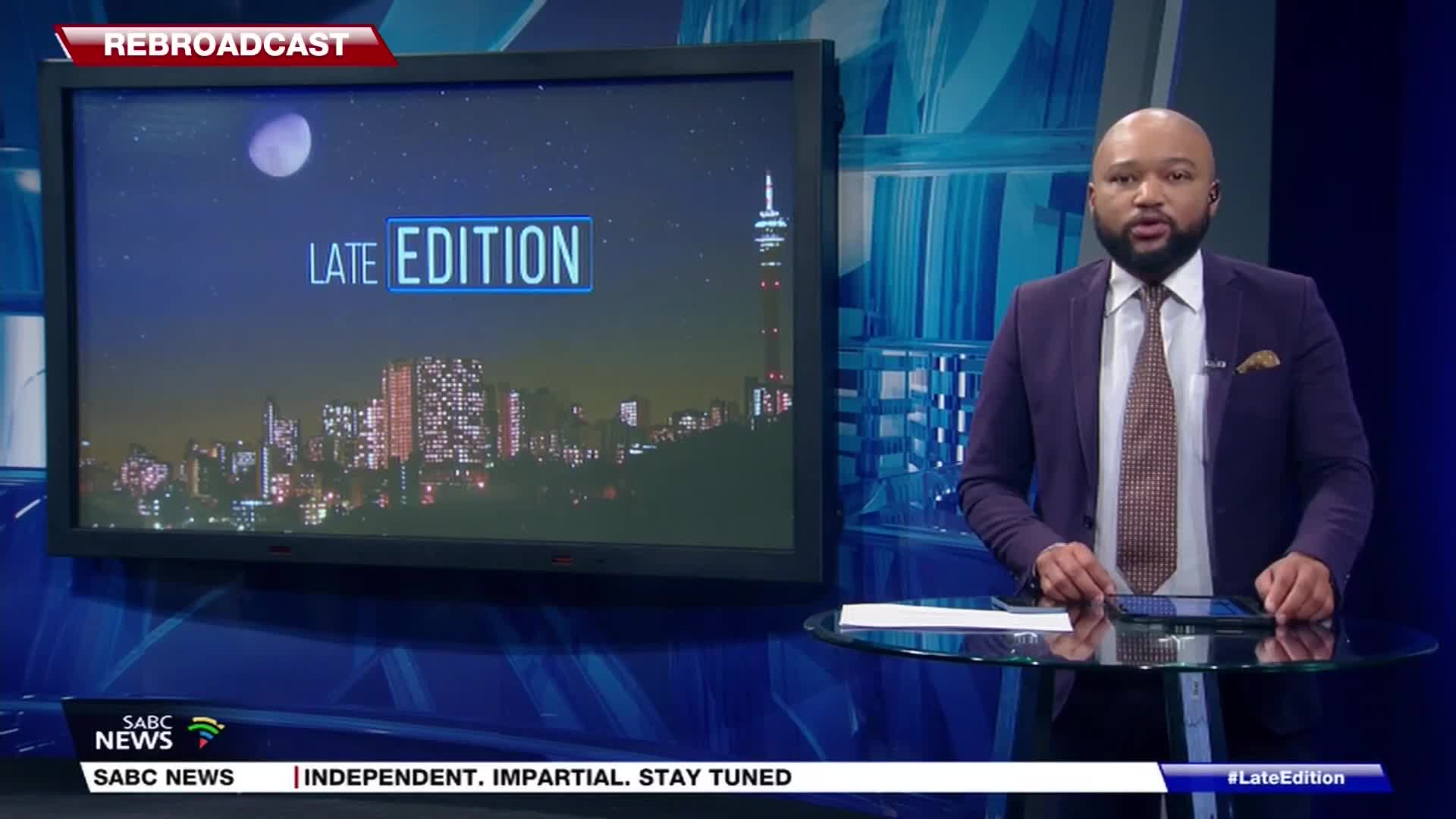 The SABC News anchor, in a purple suit, is speaking directly to the camera. Behind him, the "Late Edition" backdrop shows a cityscape with the iconic Hillbrow Tower.
