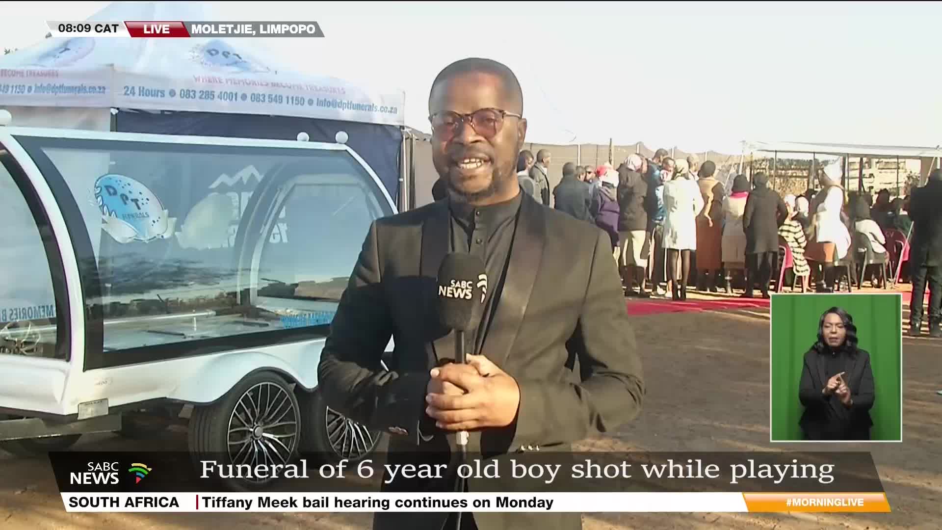 An SABC News reporter, microphone in hand, stands before a large glass-sided hearse. Behind him, mourners gather on a red carpet, awaiting the funeral in Moletjie, Limpopo, South Africa.

