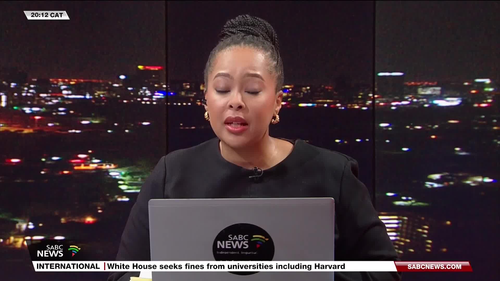 A woman in a black top speaks directly to the camera, her face illuminated by the studio lights, with the SABC News logo clearly visible. Behind her, a nighttime cityscape twinkles through a large window.
