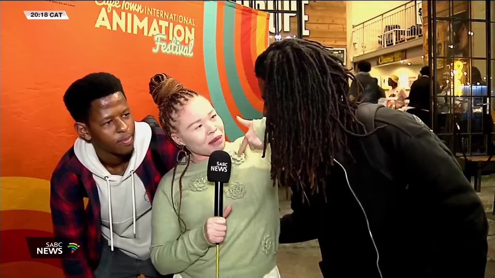 An SABC News reporter, holding a microphone, gestures towards a man with dreadlocks. Beside her, another man in a plaid shirt looks on, as the Cape Town International Animation Festival backdrop looms behind them.
