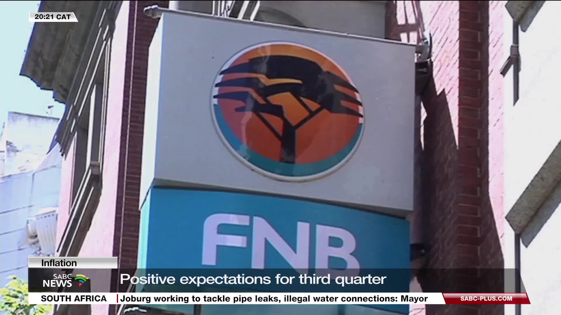 The FNB sign, with its orange and black tree logo, is mounted on a building, casting a long shadow. SABC News is reporting on positive expectations for the third quarter.
