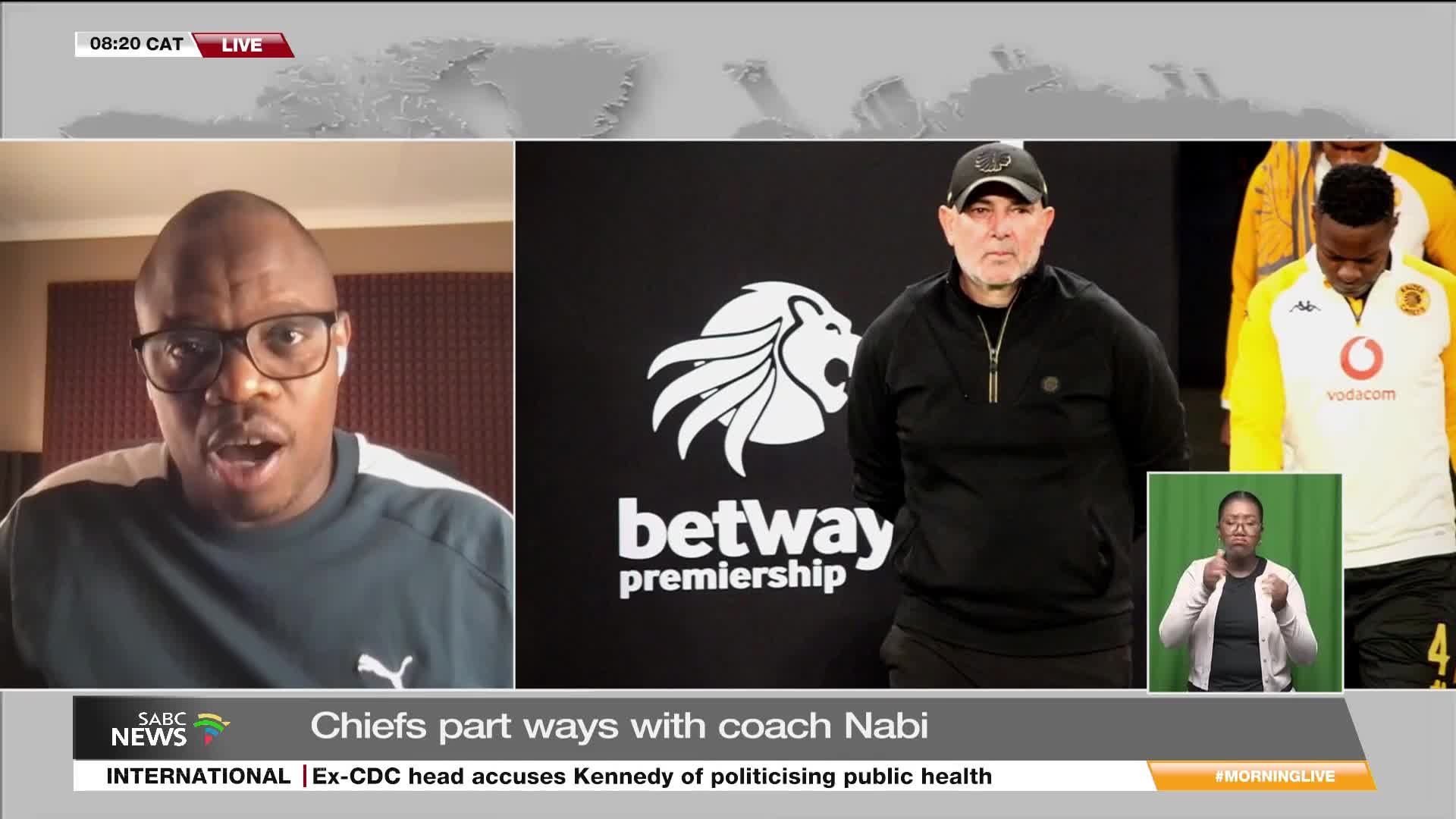 A man in glasses speaks intently, his face filling the left side of the screen on SABC News. To the right, a graphic shows a coach with the Betway Premiership logo and a sign language interpreter.

