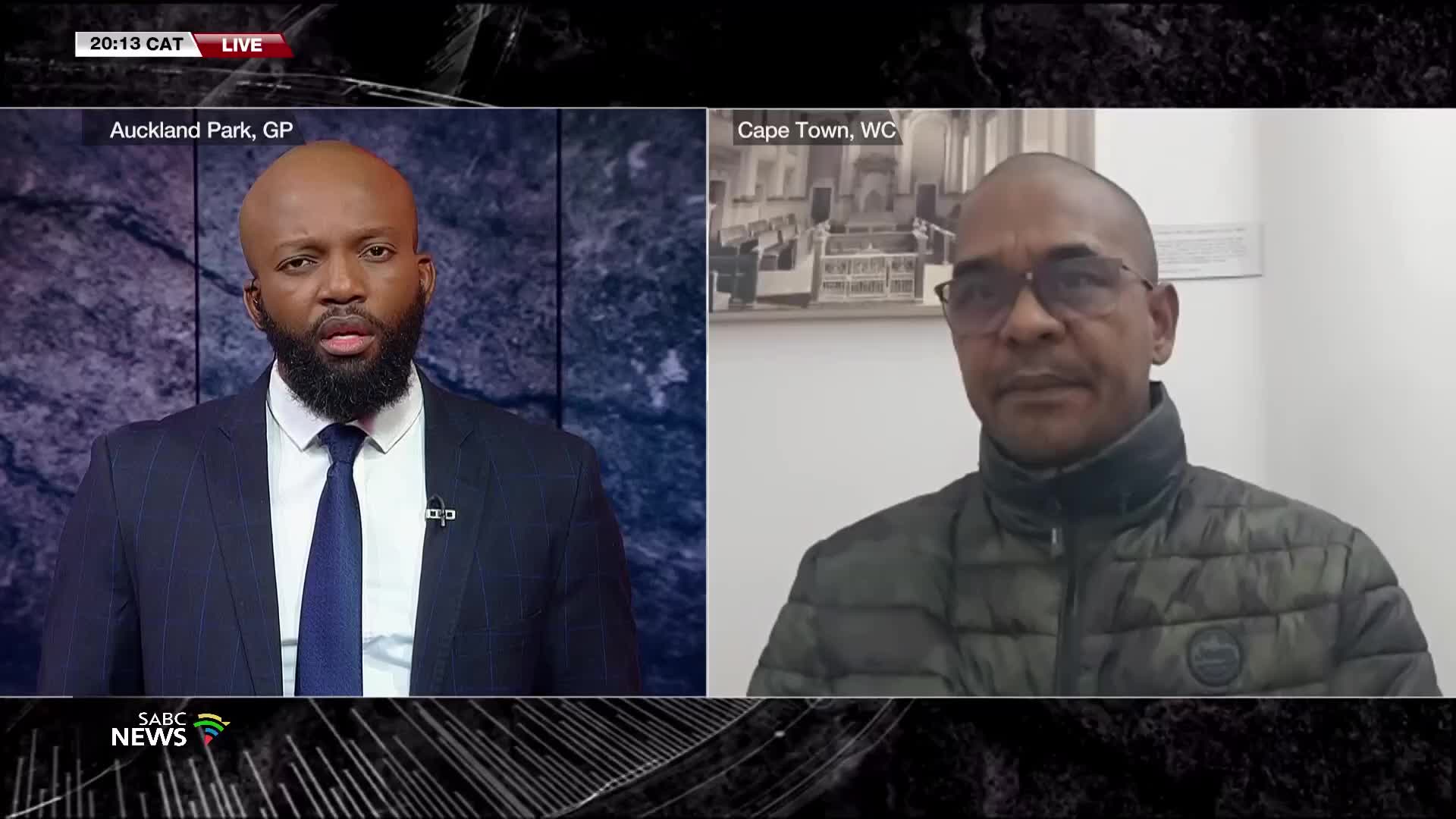 On SABC News, a man in a suit, broadcasting from Auckland Park, is speaking.  Across the screen, a man in a jacket from Cape Town is listening intently, framed against a picture on the wall.
