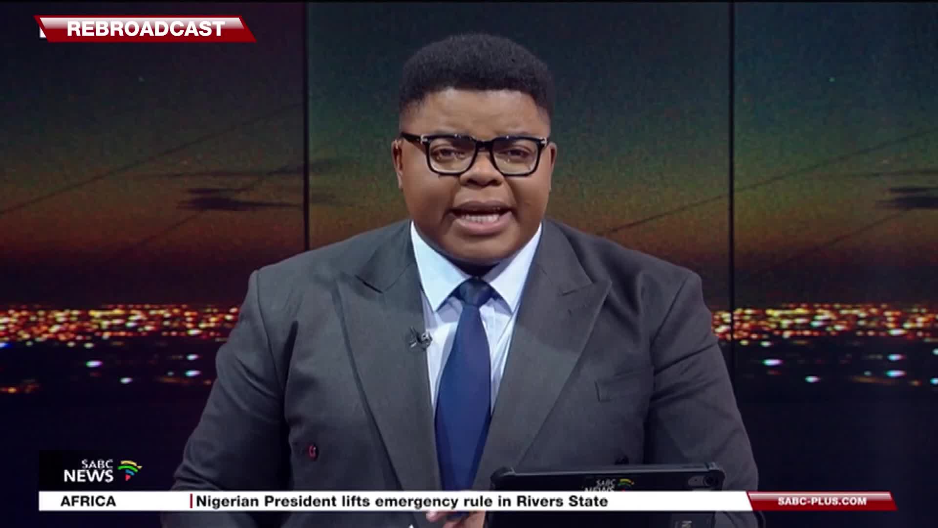 The SABC News anchor, wearing a suit and glasses, is speaking directly to the camera. Below, a headline about Nigeria scrolls across the bottom of the screen.
