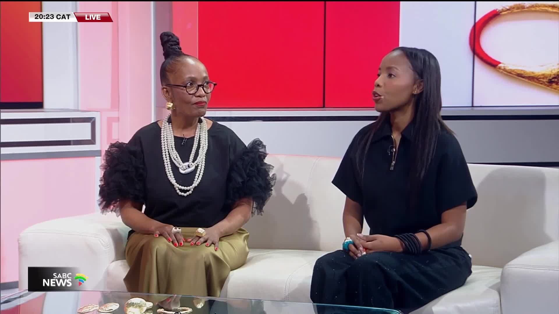 On the SABC News set, two women sit on a white couch, one with a high bun and pearls, the other in a black dress, speaking. The "LIVE" indicator glows above them, as the broadcast continues.
