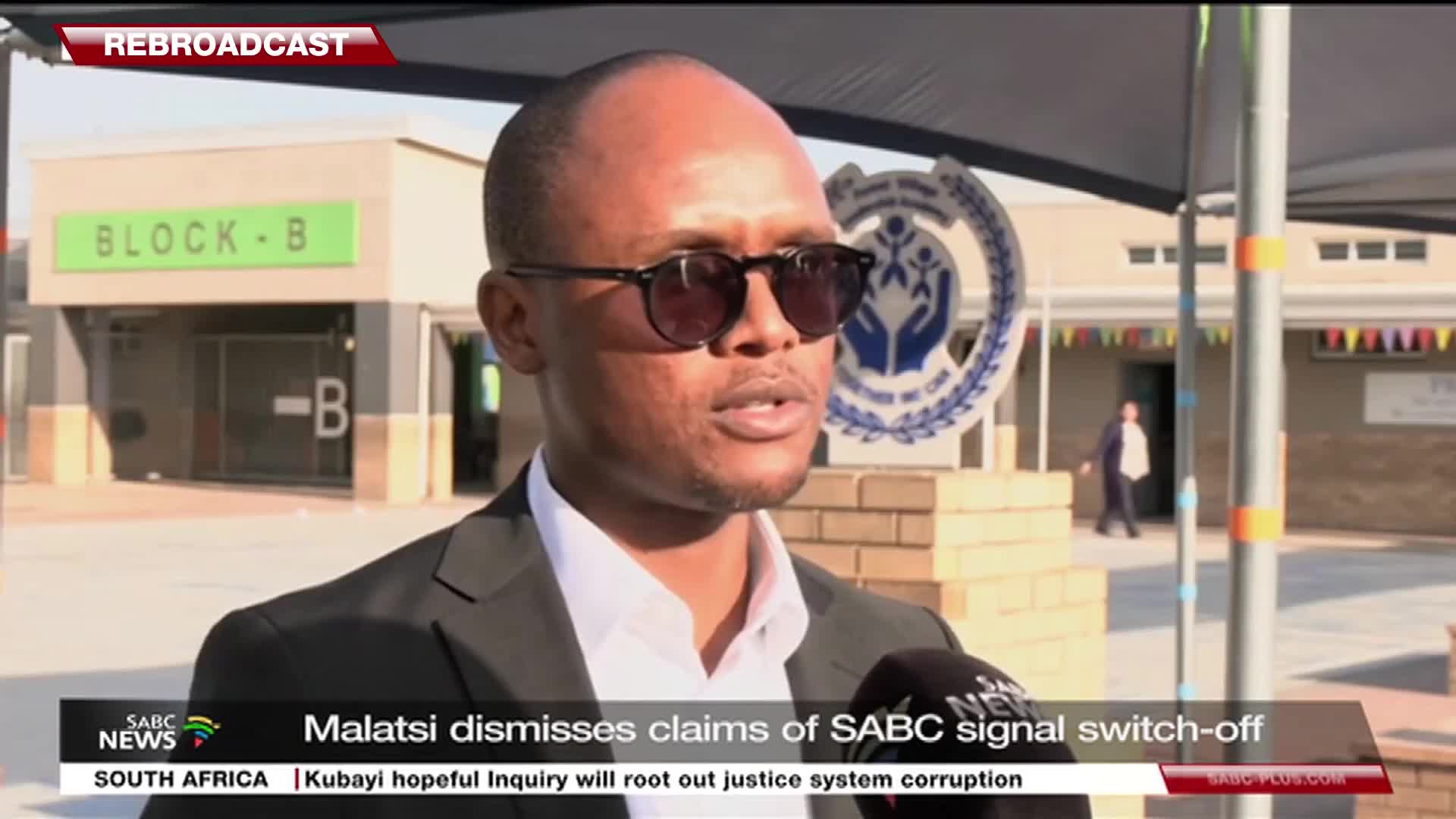 A man in sunglasses speaks into a microphone, the SABC News logo visible below. Behind him, a building with "BLOCK-B" painted on it stands in the sunlight.
