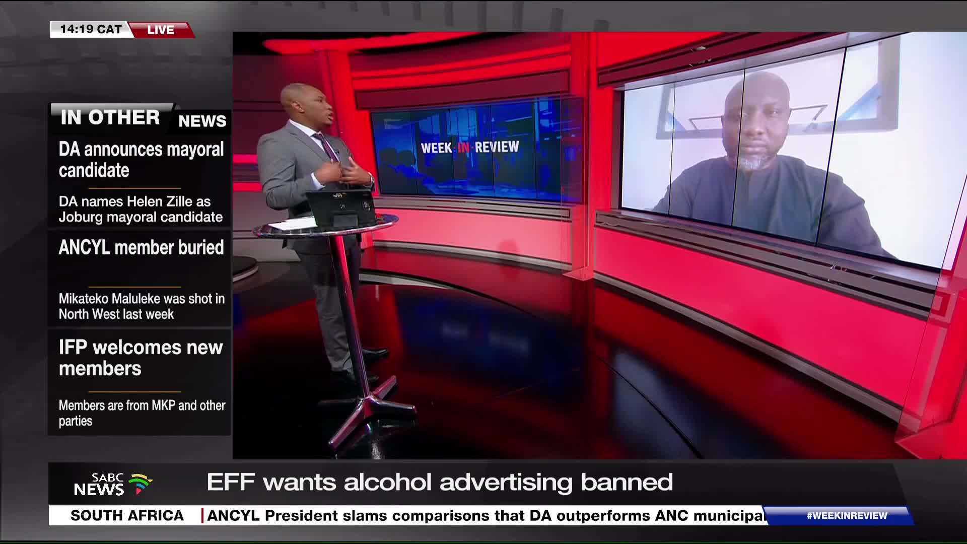 A man in a suit gestures while standing behind a small podium, likely on SABC News in South Africa. Across the studio, a screen shows another man, also on camera, in what appears to be a video call.
