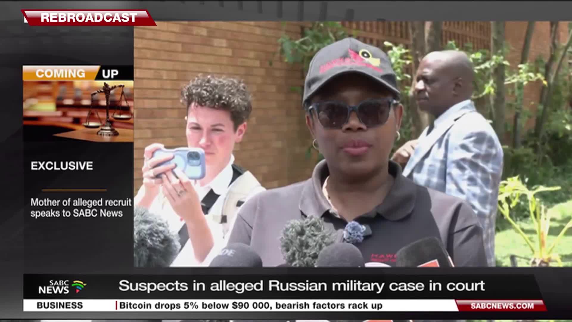 A woman in sunglasses and a cap speaks into a cluster of microphones, likely for SABC News. Behind her, another person holds up a phone, filming the scene.
A woman in sunglasses and a cap speaks into a cluster of microphones, likely for SABC News. Behind her, another person holds up a phone, filming the scene.