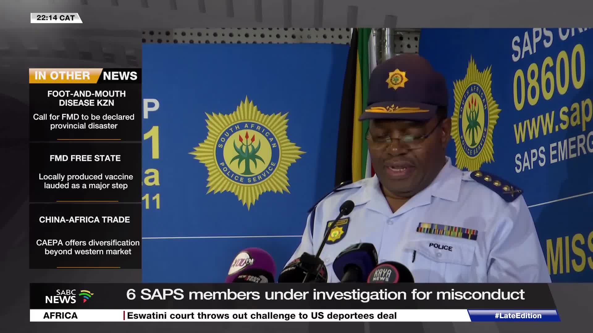 A police officer in uniform speaks at a podium, flanked by microphones. Behind him, a blue banner displays the South African Police Service emblem.