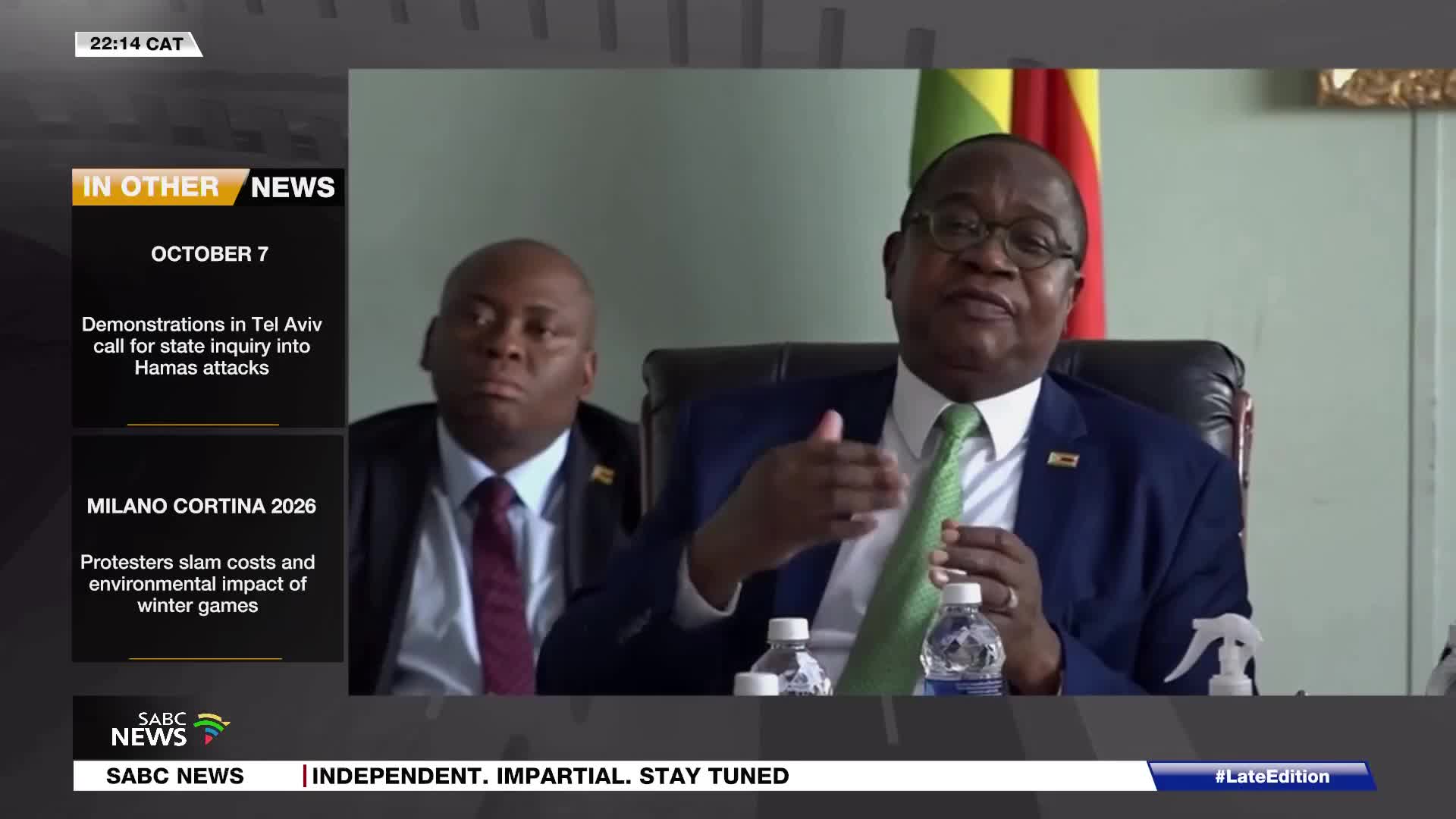A man in a blue suit and green tie gestures with his right hand while speaking. Beside him, another man in a dark suit and maroon tie looks on intently. The SABC News logo appears at the bottom left of the screen.