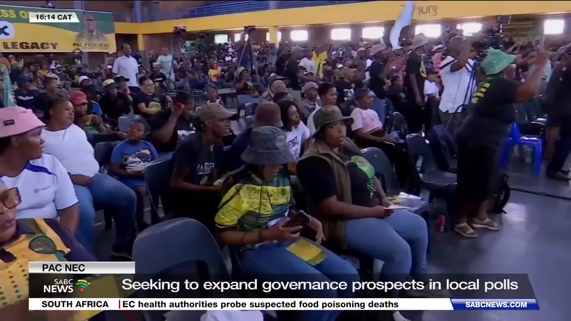 A woman in a green hat raises her hand, her body angled towards the stage, while a crowd of people sits in chairs, many looking towards the front. SABC News is broadcasting this gathering, likely a political event in South Africa, with a banner in the background mentioning "PAC NEC" and "Legacy."
