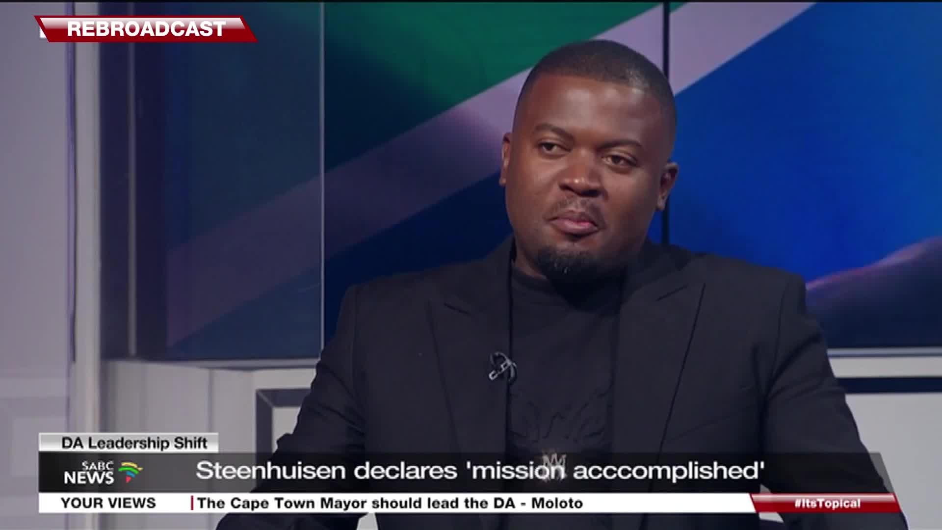 A man in a black suit sits in a studio, the SABC News logo visible below him. He looks towards the camera, a subtle expression on his face.