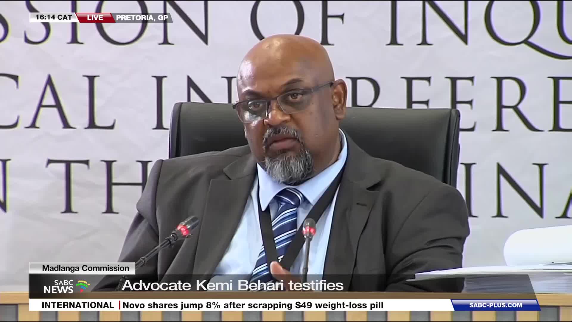 Advocate Kemi Behari is speaking at the Madlanga Commission. The SABC News broadcast shows him seated, wearing a dark suit and tie.