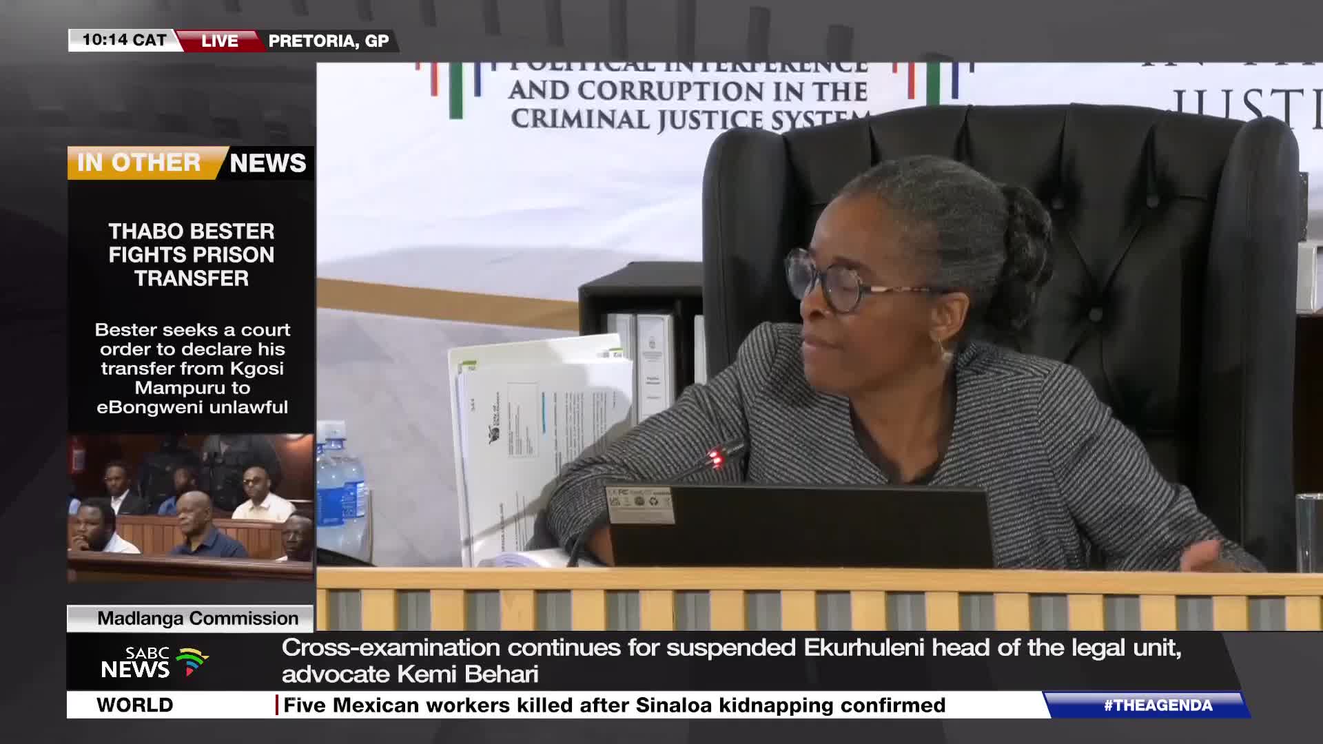 A woman in glasses, with her hair pulled back, leans forward at a desk in a formal setting. On the screen behind her, the words "POLITICAL INTERFERENCE AND CORRUPTION IN THE CRIMINAL JUSTICE SYSTEM" are displayed.