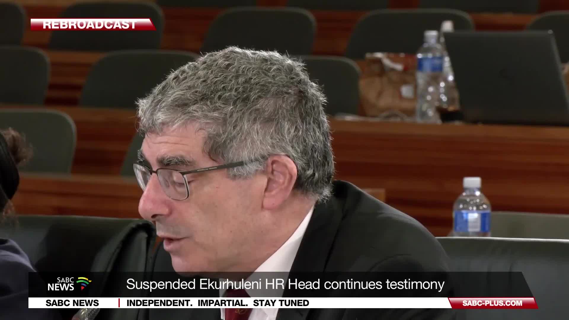 A man with grey hair and glasses speaks, his mouth slightly open. The SABC News banner at the bottom of the screen identifies him as the suspended Ekurhuleni HR Head continuing his testimony. A man with grey hair and glasses speaks, his mouth slightly open. The SABC News banner at the bottom of the screen identifies him as the suspended Ekurhuleni HR Head continuing his testimony.