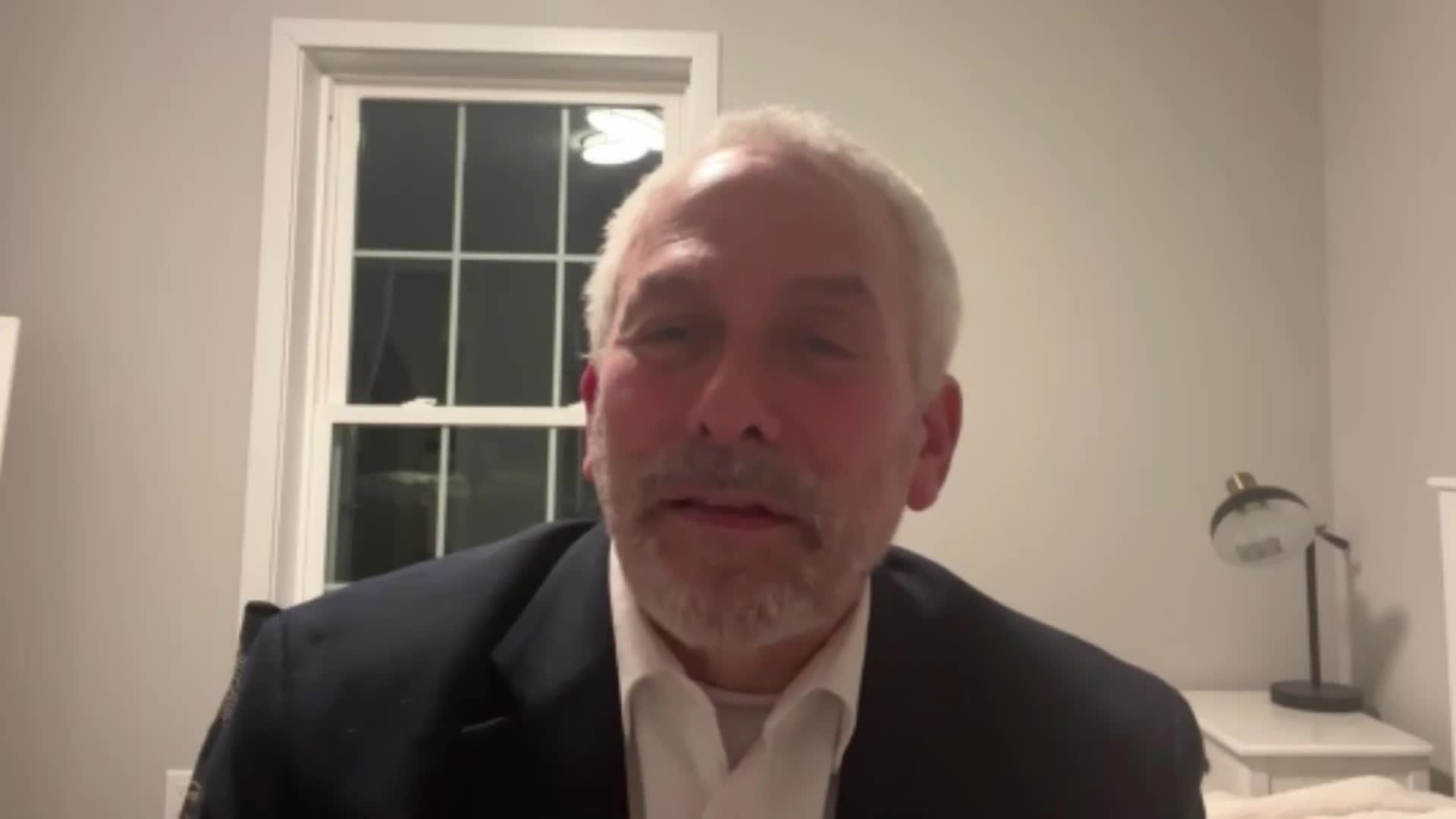 A man with grey hair and a beard, wearing a dark suit jacket over a white shirt, speaks directly to the camera. Behind him, a window with multiple panes is visible, and a desk lamp sits on a nightstand. A man with grey hair and a beard, wearing a dark suit jacket over a white shirt, speaks directly to the camera. Behind him, a window with multiple panes is visible, and a desk lamp sits on a nightstand.