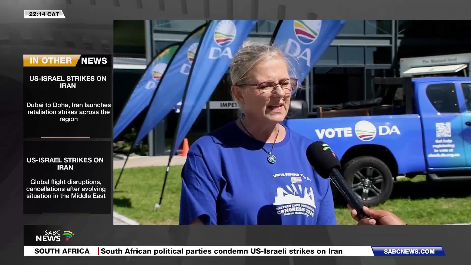 A woman in a blue t-shirt speaks into a microphone, with a blue pickup truck and DA party flags behind her. The SABC News chyron indicates this is a report from South Africa. A woman in a blue t-shirt speaks into a microphone, with a blue pickup truck and DA party flags behind her. The SABC News chyron indicates this is a report from South Africa.