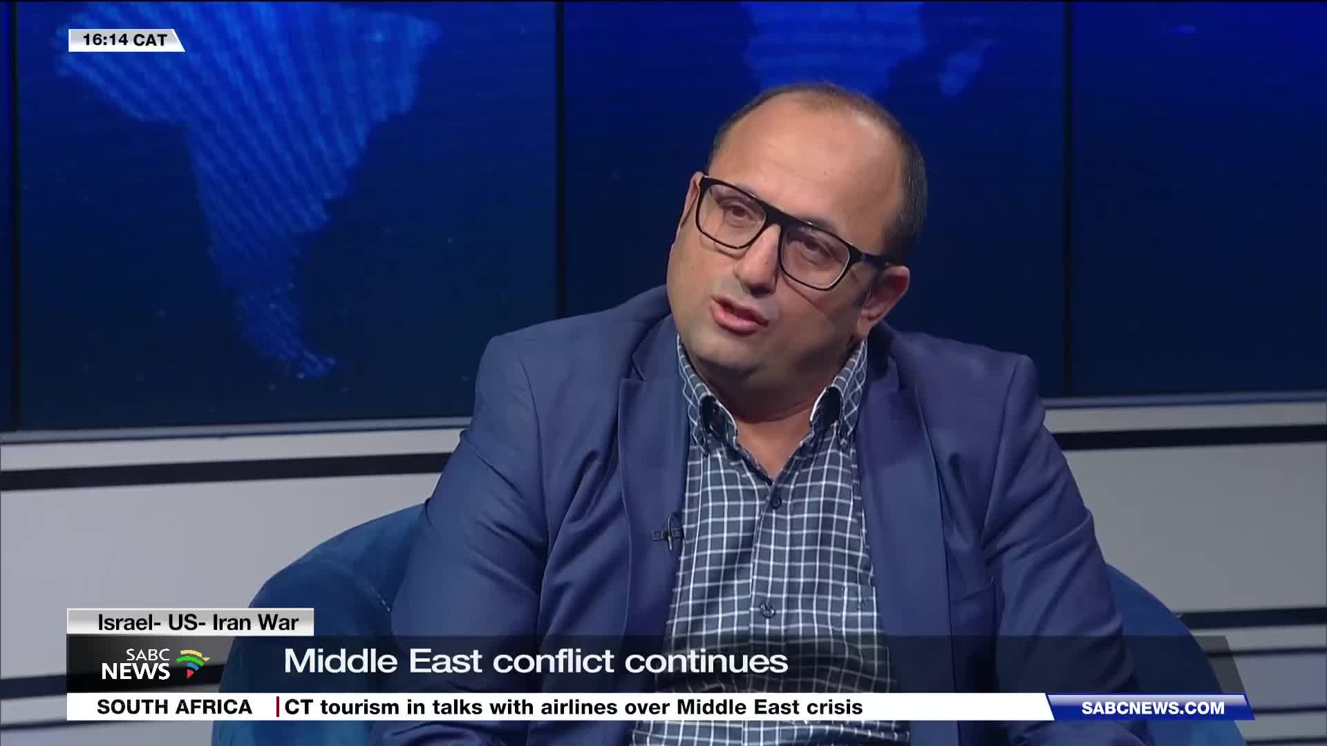 A man with glasses is speaking, his blue blazer open over a checkered shirt. A graphic on the screen behind him states "Middle East conflict continues" and "SABC NEWS." A man with glasses is speaking, his blue blazer open over a checkered shirt. A graphic on the screen behind him states "Middle East conflict continues" and "SABC NEWS."