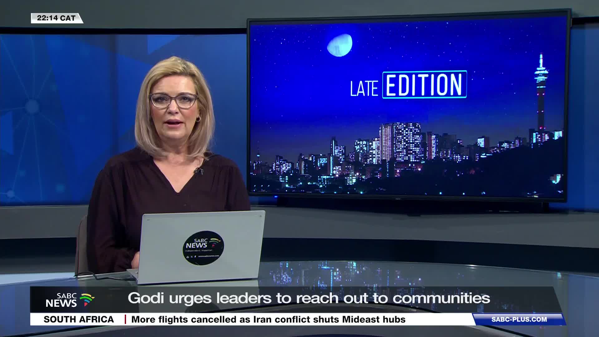 The SABC News anchor speaks directly to the camera, her words about Godi urging leaders to reach out appearing on a ticker below. Behind her, a nighttime cityscape with a prominent tower fills the screen. The SABC News anchor speaks directly to the camera, her words about Godi urging leaders to reach out appearing on a ticker below. Behind her, a nighttime cityscape with a prominent tower fills the screen.