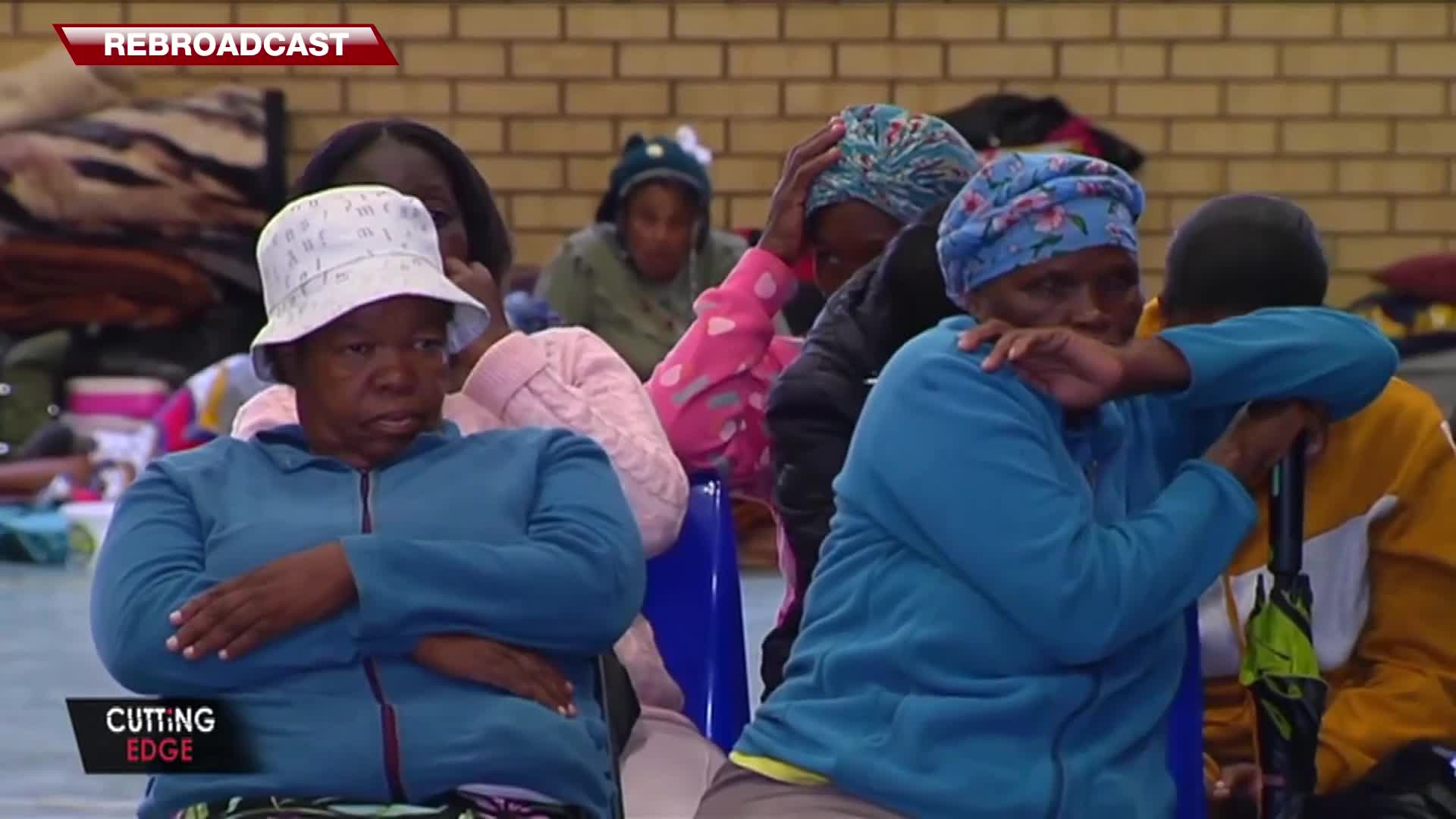 A woman in a blue jacket with her arms crossed gazes forward, her expression somber. Beside her, another woman in a similar blue top leans in, her arm around someone's shoulders, holding a folded umbrella. A woman in a blue jacket with her arms crossed gazes forward, her expression somber. Beside her, another woman in a similar blue top leans in, her arm around someone's shoulders, holding a folded umbrella.