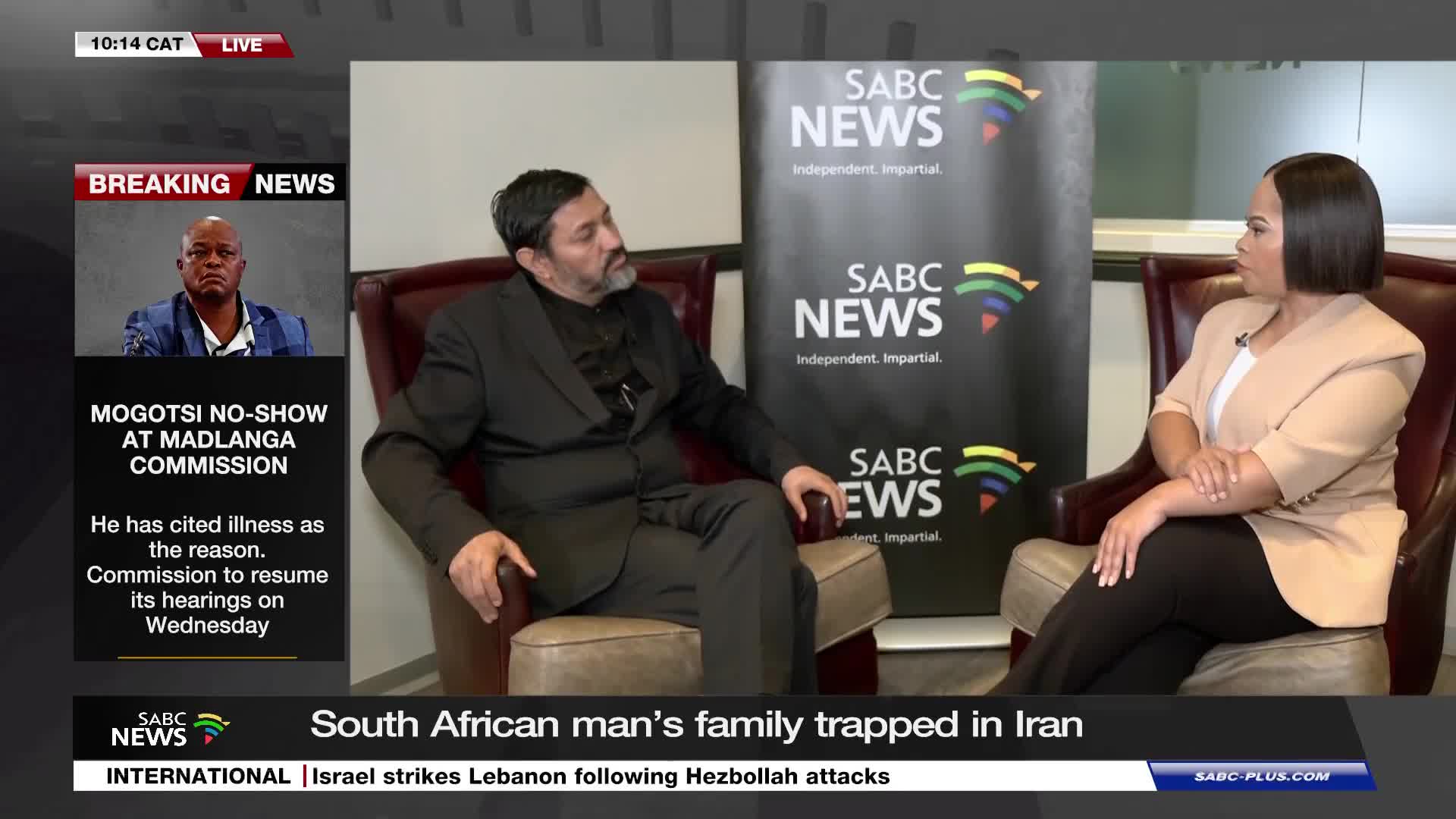 A man in a dark suit sits in an armchair, facing a woman in a tan blazer. Behind them, a SABC News banner stands tall. A man in a dark suit sits in an armchair, facing a woman in a tan blazer. Behind them, a SABC News banner stands tall.